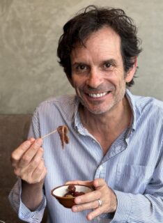 Author Chris Beckman holding an anchovy on a toothpick in one hand and a bowl in the other. He is wearing a blue button-down shirt and looking into the camera.