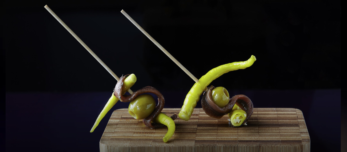 Two Gilda pintxos with a long green pepper, a glistening anchovy and a green olive skewered on a long toothpick in a wooden block, against a black background.