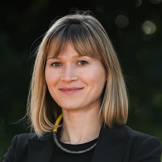 Portrait of a young woman with shoulder-length light brown hair. She is wearing a dark round-necked top and a necklace.