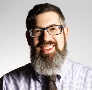 Head and shoulders portrait. A smiling man with spectacles, short gray hair and a long gray beard, wearing a blue check shirt and riddish tie.